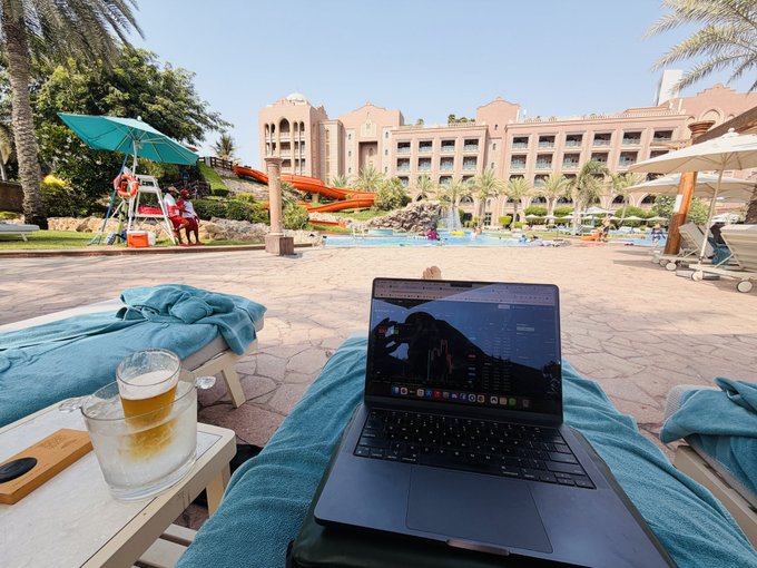 A MacBook rests on a poolside lounge chair beside a glass of beer served in its own ice bucket, with a resort featuring a red water slide and people enjoying the pool visible in the background.