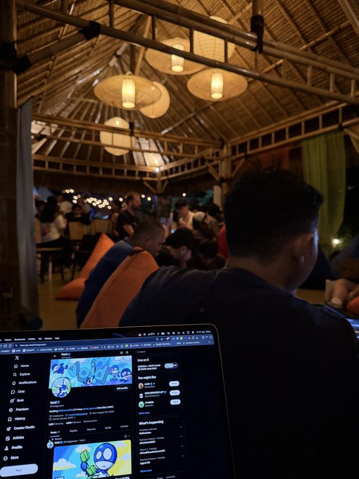 A dimly lit, rustic open-air venue with thatched roofing and paper lanterns, where a person in the foreground uses a laptop displaying the Twitter/X interface while others socialize in the background.