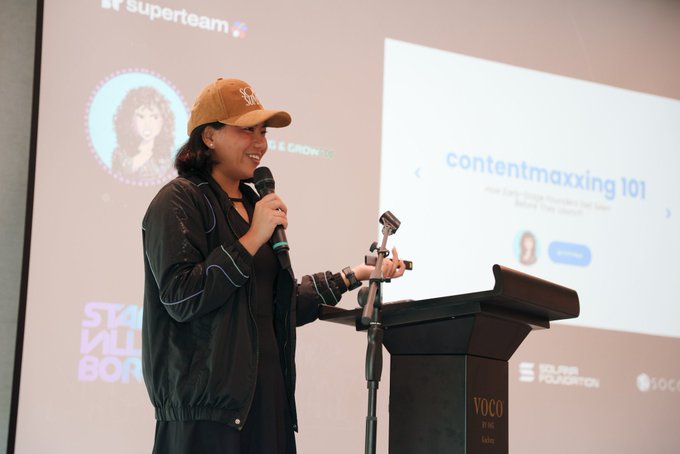 A woman wearing a Superteam cap speaks at a podium with a microphone, presenting a slide titled 'contentmaxxing 101' during what appears to be a Solana Foundation event.