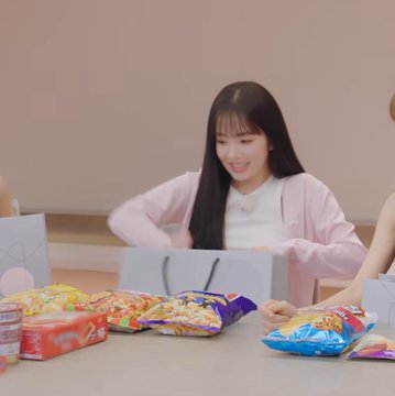 A young woman in a pink cardigan smiling while sitting at a table covered with various snack packages including chips and candy.