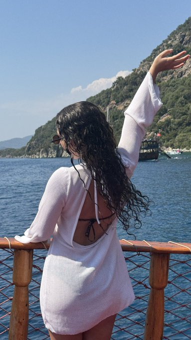 A woman with long curly hair wearing a white cover-up over a swimsuit stands on a boat, gesturing toward a scenic coastal landscape.