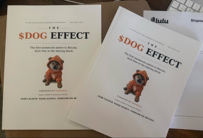 Two copies of a book titled 'THE $DOG EFFECT' - described as 'A Cryptolution Research Handbook' and 'The first memecoin native to Bitcoin, born free in the halving block,' featuring a Shiba Inu dog in an orange hoodie on the cover, with shipping materials from Lulu visible.