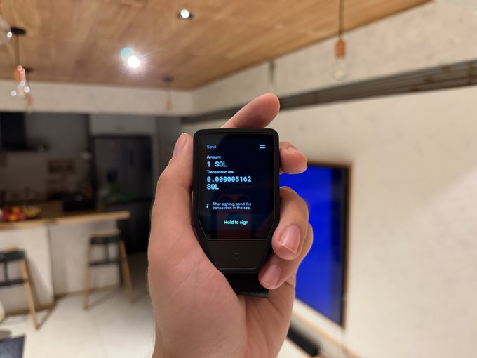 A hand holds a hardware crypto wallet displaying a transaction confirmation screen to send 1 SOL to address 6...0w885162 SOL, with a modern kitchen visible in the blurred background.