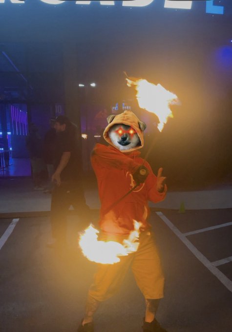 A performer in an orange hoodie and yellow pants breathes fire while holding flaming torches in both hands. They wear a mask with glowing red eyes, appearing to be a dog or animal character.