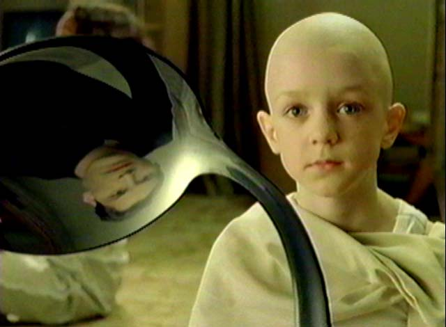 A bald child looking directly at the camera while holding a curved metallic object that reflects another person's face.