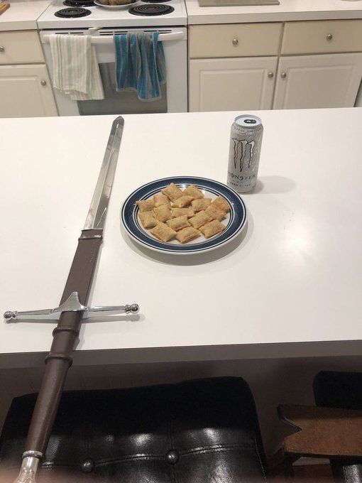 A large decorative medieval-style sword lying on a white kitchen counter next to a plate of pastries and a Monster Energy drink can.