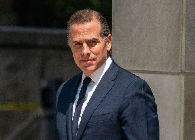 A man with slicked-back dark hair wearing a navy suit and tie stands outside a stone building, looking directly at the camera with a serious expression. The image appears to relate to the post about Hunter Biden launching a cryptocurrency meme coin, likely depicting him as the subject of the news.