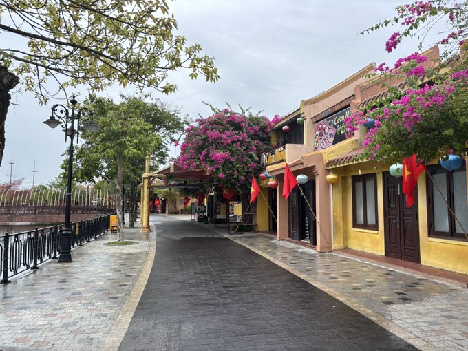 An empty street scene featuring yellow buildings with red flags and hanging lanterns, decorated with vibrant pink bougainvillea flowers under an overcast sky.