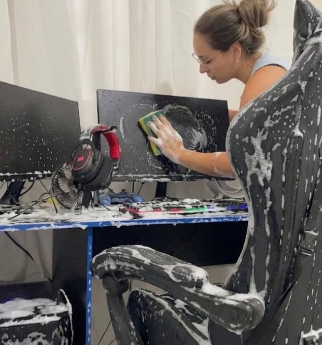 A woman wearing glasses cleans a computer monitor while covered in foam; a gaming chair, headphones, and other equipment around her are also splattered with foam.