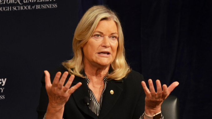 A blonde woman in a black blazer gestures with both hands while speaking at what appears to be a business school event, with partial text "UNIVERSITY" and "BUSINESS" visible on the dark backdrop behind her.