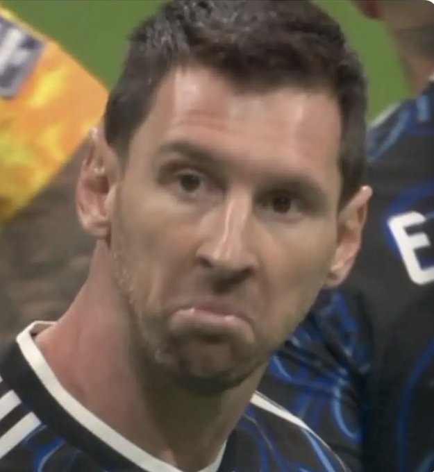 A close-up photo of soccer player Lionel Messi wearing a black and blue striped jersey, with a neutral expression and slightly pursed lips.