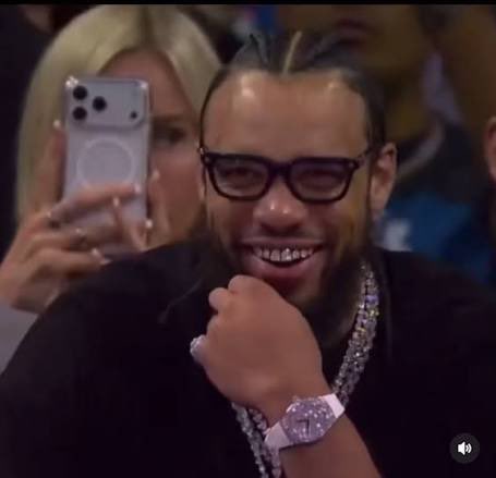 A man with braided hair, dark sunglasses, diamond grillz, a silver watch, and a chain necklace smiles at what appears to be a sporting event, with a blonde woman taking a photo in the background.