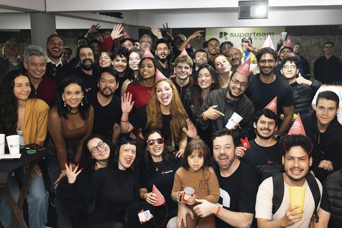 A large group photo of smiling people wearing party hats, gathered for what appears to be a 'Superteam' celebration, with one young child being held in the center.