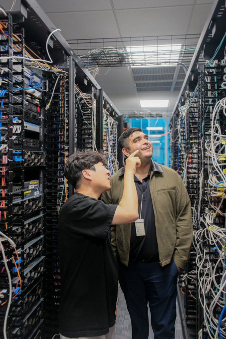 Two young men stand in a server room lined with networking racks and tangled cables, with one wearing a lanyard pointing upward.