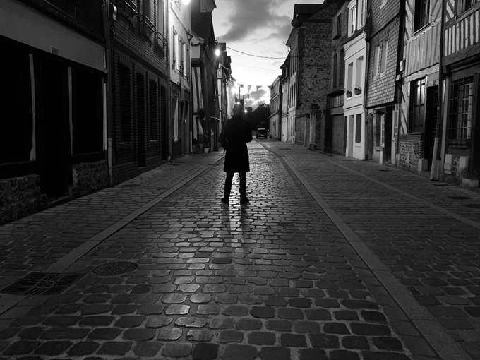A solitary figure stands alone on a dimly lit, empty cobblestone street, captured in moody black and white that emphasizes the atmospheric isolation of the scene—perfectly matching the contemplative, direction-refusing tone of the photography series.