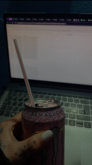 A hand holding a pink energy drink can with a straw, featuring Portuguese text 'SEM AÇÚCAR', positioned in front of an open laptop screen.