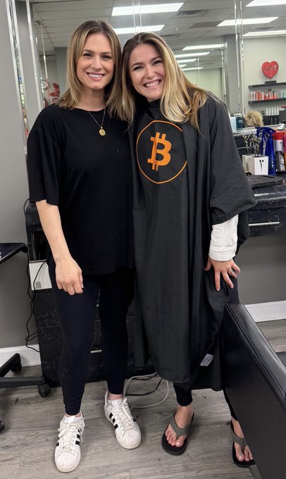 The image features two women standing together in a hair salon. They are both wearing black tops and have their arms around each other's shoulders. One of them has a large "bitcoin" t-shirt on, while the other wears a cape that says "bitcoin." The woman with the "bitcoin" t-shirt is smiling at the camera, and they appear to be posing for a photo together.