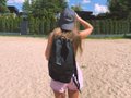A small image of a woman with long hair, wearing a cap and a backpack, standing on a sandy surface with a fence in the background.