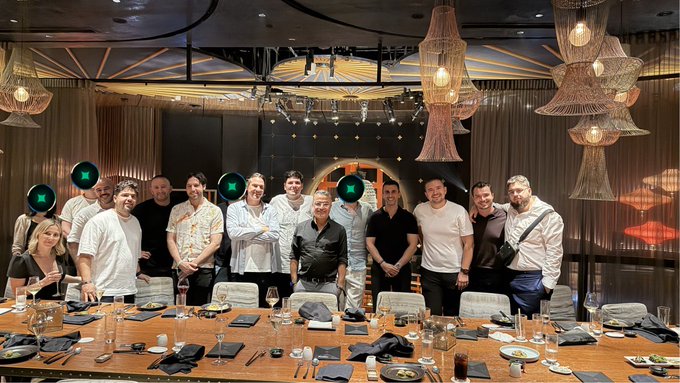 A group of people pose together behind a long dining table set with glasses and place settings in an upscale restaurant with ornate chandeliers.