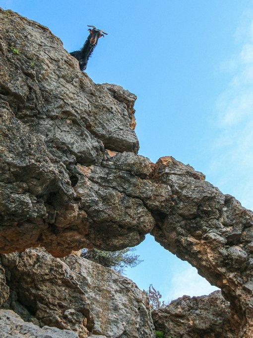 A low-angle photograph of a rugged rocky cliff or natural stone arch against a blue sky, with a person dressed in dark clothing standing at the top edge looking down.