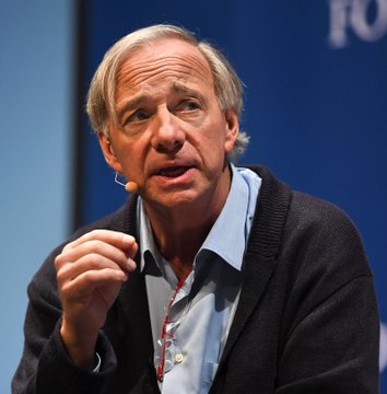 An older man with gray hair, wearing a light blue collared shirt and dark cardigan, gestures with his hand while speaking into an earpiece microphone against a blue backdrop featuring partial "FO" text. The image likely depicts Ray Dalio, the billionaire investor referenced in the caption discussing his small Bitcoin allocation as a store of value akin to gold.