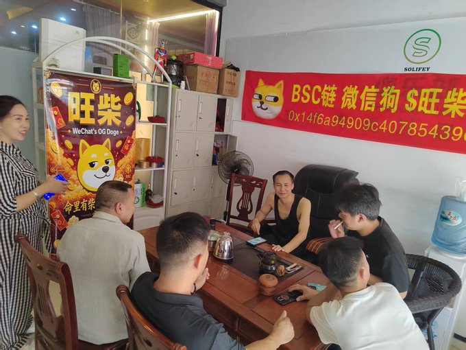 A group gathers around a table beneath banners advertising "旺柴" (WeChat's OG Doge) and a BSC wallet address (0x14f6a94909c40785439...), with the SOLIFEY logo visible above, as the community has taken down the Wangchai meme banner to focus on serious crypto business.