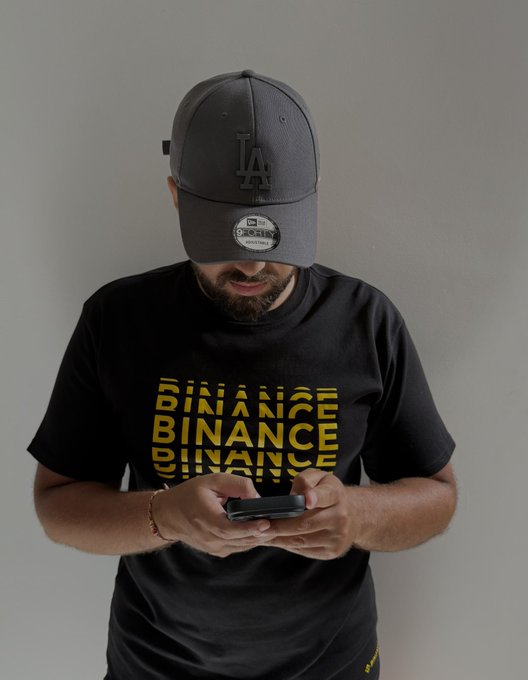 A man wearing a dark grey LA baseball cap and a black t-shirt with repeated yellow 'BINANCE' text looks down at his phone.