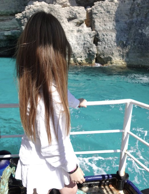 A person with long brown hair, wearing a white outfit, stands on a boat looking out at bright turquoise water and rocky cliffs.