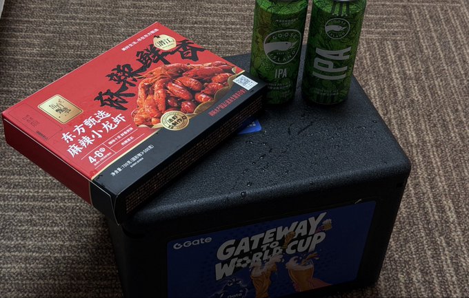 A red Chinese food package labeled '麻辣小龙虾' (Spicy Crayfish) sitting on a black 'Gateway World Cup' branded box, accompanied by two green cans of what appears to be Green Lake IPA beer on a carpeted floor.