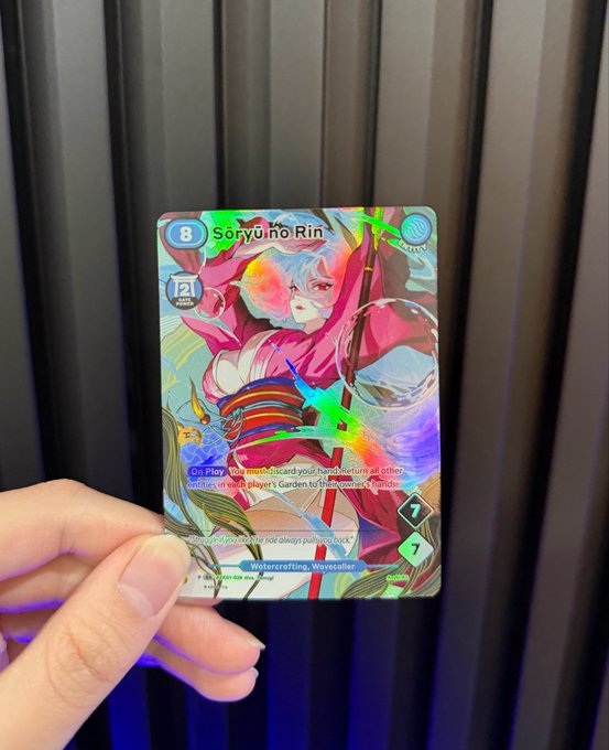 The image shows a person holding up a card with a picture of a woman, possibly a character from a video game or anime. The card has a blue background and features a pink and white design that includes a sword and a shield. The text on the card reads "Sery Uno Rin".