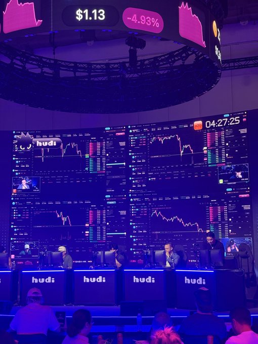 A crypto trading event scene with people working at computer stations in front of large screens displaying price charts and graphs under purple lighting, with a ticker showing '$1.13 -4.93%' and a countdown timer reading '04:27:25'.