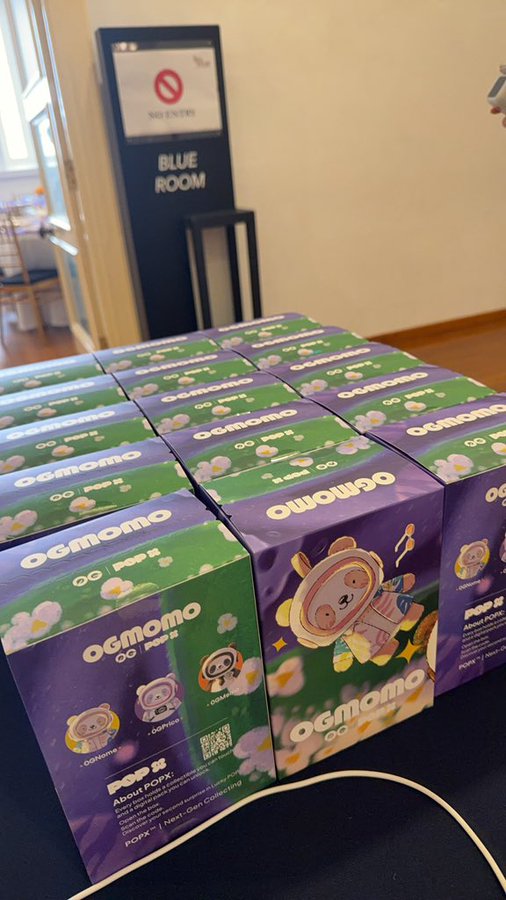 A collection of POP MART 'OGmomo' collectible figure boxes in purple and green packaging arranged on a table near a 'Blue Room' sign.