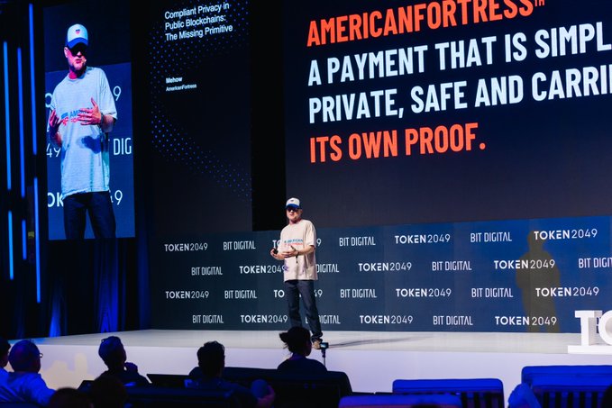 A speaker on stage at the TOKEN2049 BIT DIGITAL event presenting 'AMERICANFORTRESS' with the tagline 'A PAYMENT THAT IS SIMPLE, PRIVATE, SAFE AND CARRIES ITS OWN PROOF.'