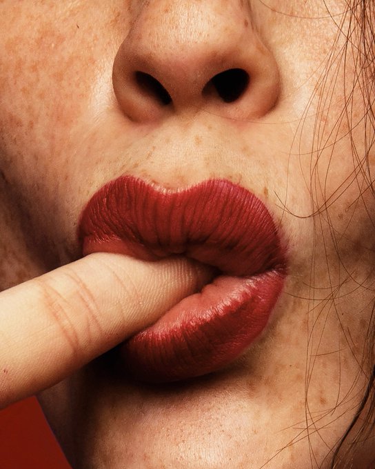 An extreme close-up photograph shows a woman's lower face with red lipstick, freckles, and a finger touching her lips.