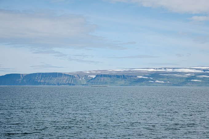 A scenic landscape photograph shows a calm blue sea in the foreground with snow-capped mountains and cliffs along the horizon under a partly cloudy sky.