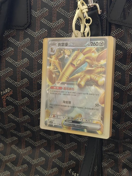 A Pokémon trading card featuring Gholdengo ex with 260 HP is displayed inside a protective case attached as a keychain to a Goyard-patterned bag.