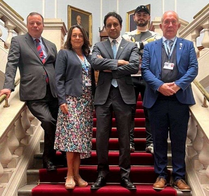 A group of five people, including one in a security uniform, pose on a red-carpeted staircase in what appears to be a government building. The image likely satirizes Elon Musk's DOGE initiative by framing his associates as a chaotic "circus," with Musk (center) standing prominently among his team members in formal attire.