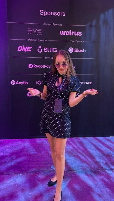 A woman wearing sunglasses, a dark polka-dot dress, and a lanyard poses in front of a sponsor board displaying logos including EVE Frontier, Walrus, ONE, SUIG, Slush, RedotPay, and Anyflo.