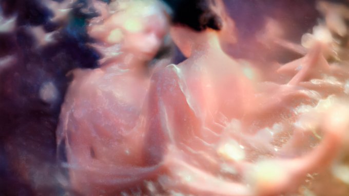 A blurry, abstract close-up image featuring soft pink, white, and purple amorphous shapes that resemble liquid, smoke, or perhaps a microscopic biological structure.