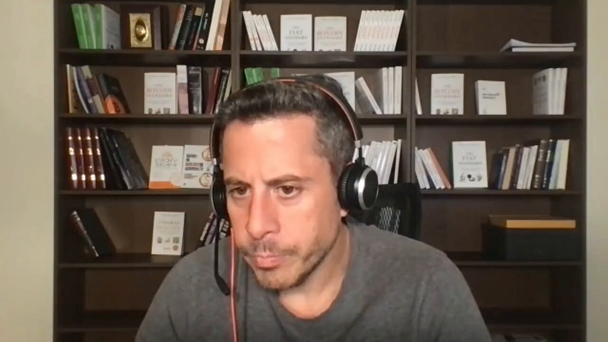 The image shows a man wearing headphones, sitting at a desk with his eyes closed while looking down at something. The background features a bookshelf filled with numerous books of various sizes. There are also two small golden objects near the top left corner of the bookshelf. The man's position and the presence of the bookshelf suggest that he might be engaged in some form of online learning or research, possibly related to the post about voting harder or Bitcoin.