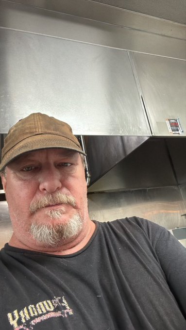 Selfie of a man wearing a brown cap and black t-shirt with 'Dynavex' text, standing in front of a stainless steel commercial vent hood.