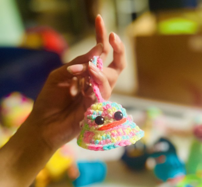 A close-up of a hand holding up a small, colorful crocheted amigurumi charm in pastel rainbow colors with googly eyes and a cute smile.