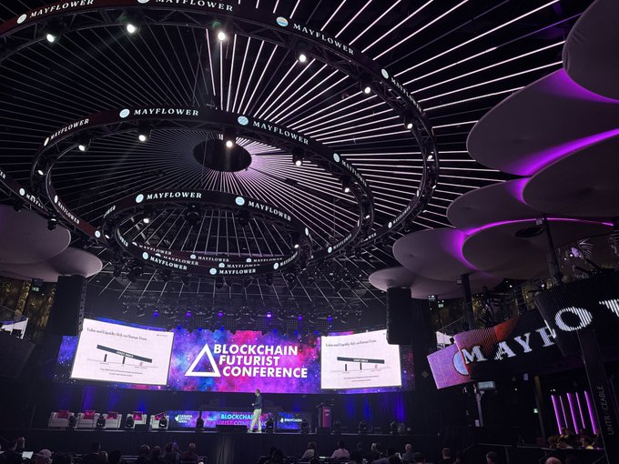 The image shows a presentation at the Blockchain Futurist Conference, with the stage lit by "Mayflower" branding, which also appears on an overhead circular structure that could be described as rings. The "rings" in the post likely refer to the circular, illuminated structures above the stage displaying the "MAYFLOWER" logo.