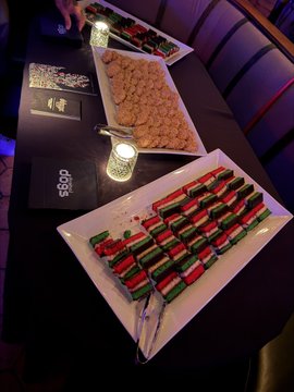A table at an event displays platters of food including what appears to be fried items and stuffed grape/pepper rolls, with '1969' printed on the black table covering and decorative lanterns.