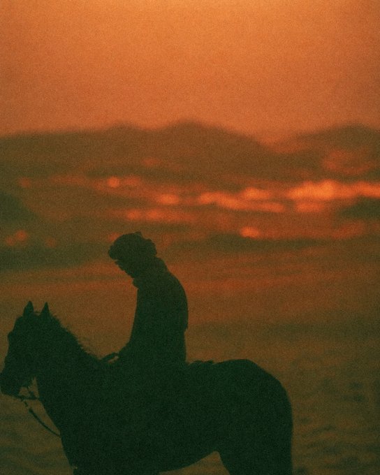 A silhouetted figure wearing a headwrap sits on horseback against a dramatic orange and red sunset sky.