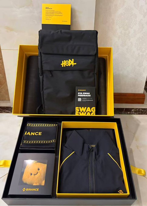A Binance branded swag box containing a navy backpack, polo shirt, and accessories with yellow 'SWAG' themed packaging.
