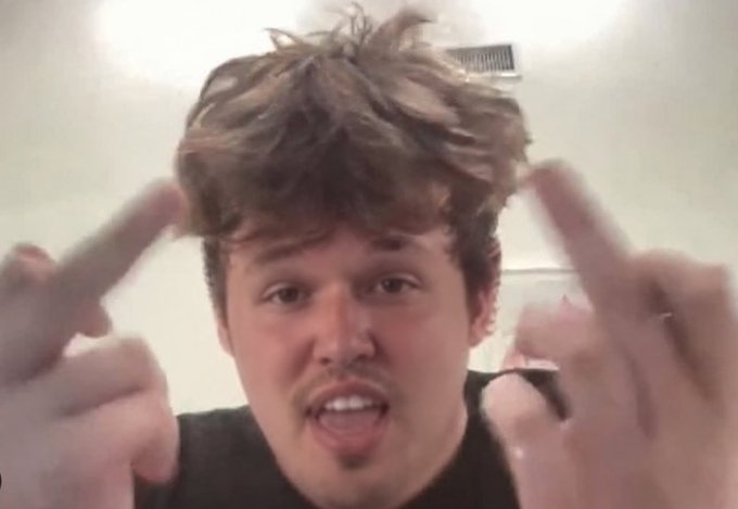 A close-up of a young man with messy brown hair and a small mustache speaking directly to the camera with both hands raised.