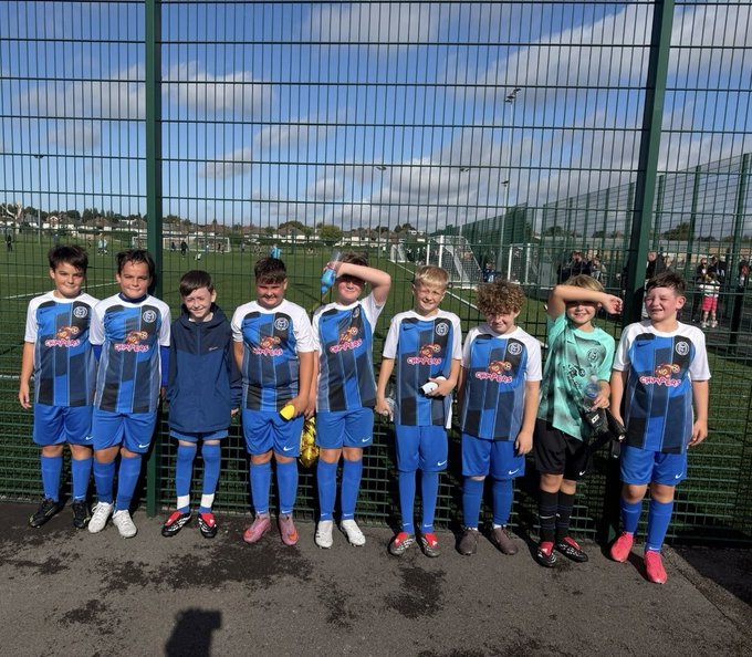 A youth soccer team poses in their blue and white uniforms in front of a fenced pitch.