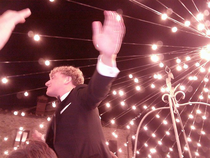 A young man in a suit waves energetically with one arm raised under a canopy of sparkling string lights, suggesting a celebratory event or performance.