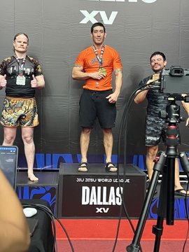Three men pose on a podium for the IBJJF Jiu-Jitsu World League XV in Dallas, with the center champion wearing an orange rashguard and black shorts, flanked by two medalists, while a camera operator films the celebration—capturing the "champ shit" moment of victory.
